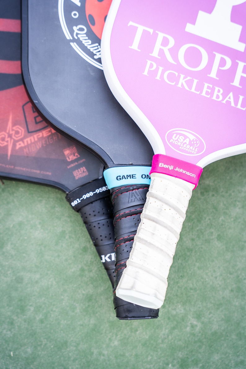 Puede incluir: Tres palas de pickleball con diferentes dise&ntilde;os y colores. La pala superior es rosa con texto blanco que dice "Trophy Pickleball". La pala del medio es negra con texto blanco que dice "Quality". La pala inferior es roja y negra con texto blanco que dice "Amped Midweight".