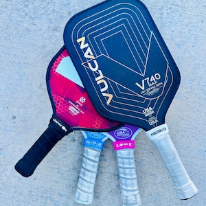 Op de afbeelding: Drie pickleball rackets met zwarte, rode en paarse vlakken. Het zwarte racket heeft gouden accenten en de tekst "VULCAN" en "V740 MAX JAY DEVILLIERS". De andere hebben witte en grijze handvatten met tekst.