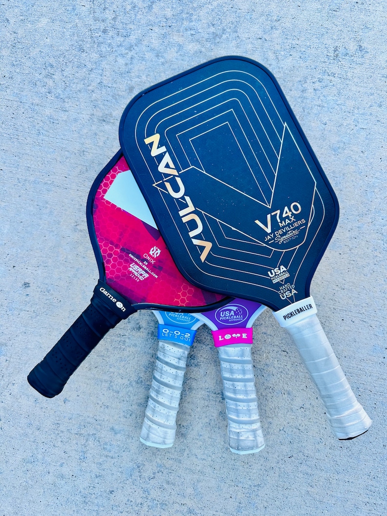 Op de afbeelding: Drie pickleball rackets met zwarte, rode en paarse vlakken. Het zwarte racket heeft gouden accenten en de tekst "VULCAN" en "V740 MAX JAY DEVILLIERS". De andere hebben witte en grijze handvatten met tekst.