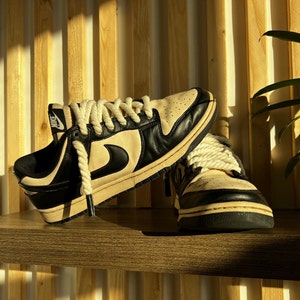 May include: A pair of black and white sneakers with white laces and a white rope tied around the laces. The sneakers are sitting on a wooden shelf.
