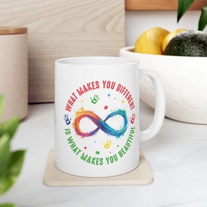 May include: White ceramic mug with a colorful infinity symbol and the text "WHAT MAKES YOU DIFFERENT IS WHAT MAKES YOU BEAUTIFUL." The design includes handprints and stars in various colors. The mug sits on a beige coaster.
