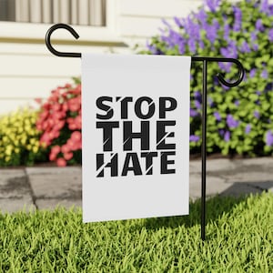May include: White garden flag with black text that reads "STOP THE HATE" in a bold, stylized font. The flag is displayed on a black metal garden stake.