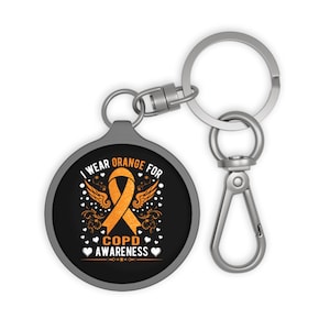 Puede incluir: Un llavero redondo negro con el texto "I wear ORANGE for COPD AWARENESS" en blanco y naranja. El diseño incluye una cinta naranja, alas y corazones. El llavero tiene un mosquetón y un llavero plateados.