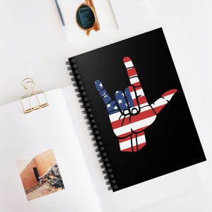 Puede incluir: Cuaderno negro con espiral con un diseño de gesto de la mano con los colores de la bandera estadounidense. El diseño incluye rayas rojas y blancas y azul con estrellas blancas. El cuaderno está abierto sobre una superficie blanca.