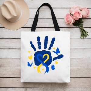 May include: A white tote bag with black handles features a blue handprint with a yellow and blue ribbon in the shape of a heart.  The design is surrounded by small blue butterflies and hearts.