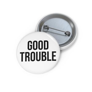 May include: Two white round buttons with the words "GOOD TROUBLE" in bold black letters. One button is in front of another button. The second button has a silver pin on the back.