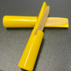 May include: A bright yellow plastic reed case, open to reveal a reed. The case is cylindrical with a rectangular lid. The reed is a light tan color. The case is approximately 5 inches long.