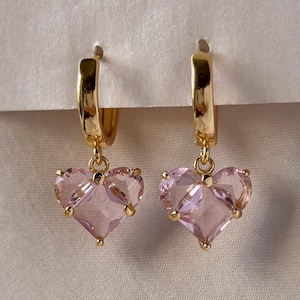 May include: A pair of gold hoop earrings featuring dangling, faceted, light pink heart-shaped charms. The earrings are suspended from a white card, showcasing a polished metallic finish on the hoops and clear, translucent pink stones.