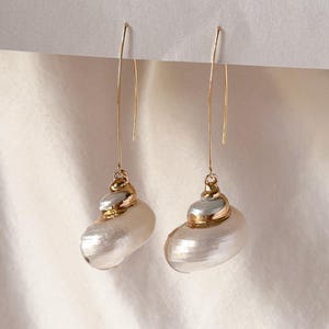 Puede incluir: Un par de pendientes colgantes con conchas marinas naturales en espiral de color blanco con acabado nacarado. Cada concha tiene un detalle de metal dorado pulido y cuelga de un largo gancho de alambre curvado de tono dorado.