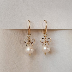 A pair of gold earrings with white pearls and bows hanging from the ear hooks.