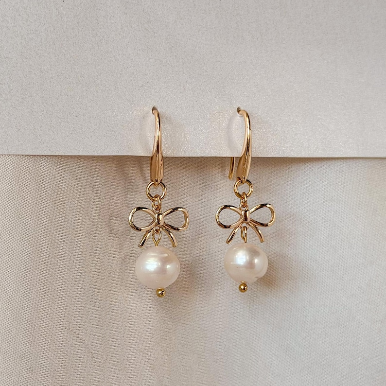 A pair of gold earrings with white pearls and bows hanging from the ear hooks.