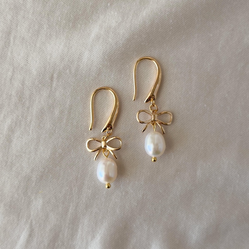 A pair of gold earrings with white pearls and bows hanging from the ear hooks.