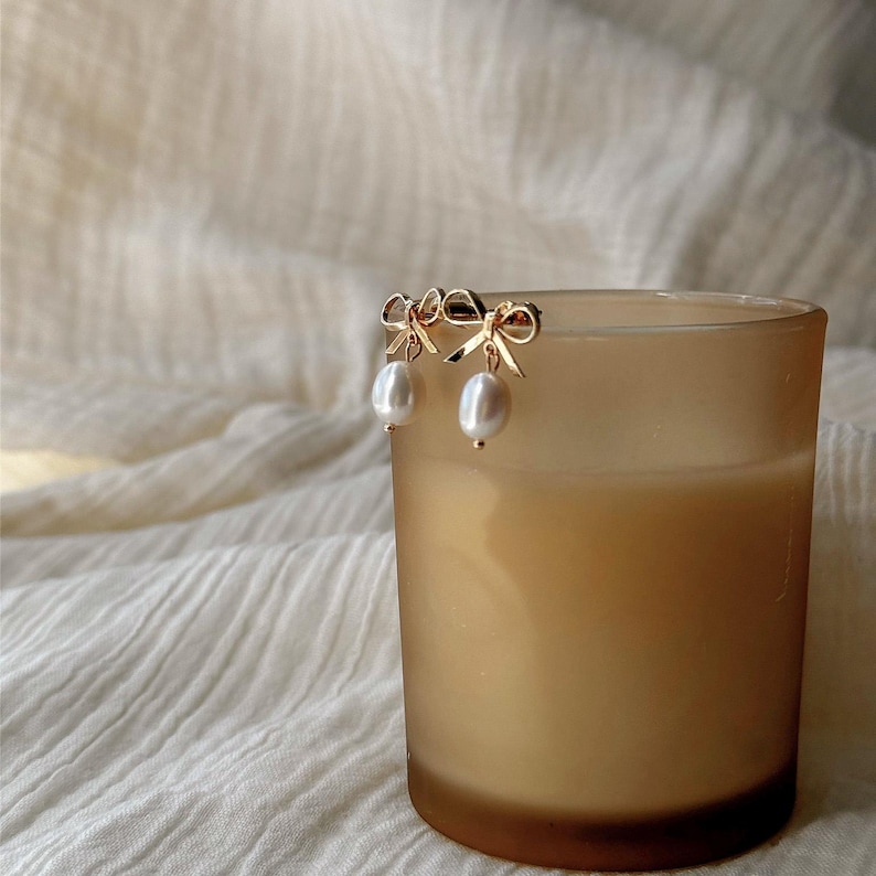 A pair of gold earrings with pearls rests on a beige candle in a glass container.