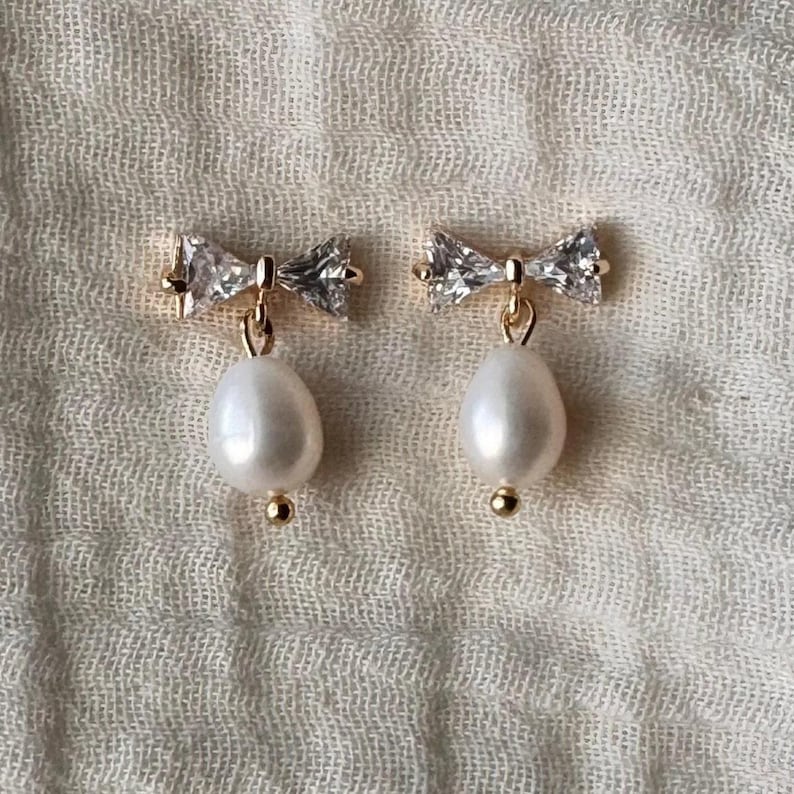 A pair of pearl drop earrings with crystal accents, displayed on a textured fabric background.