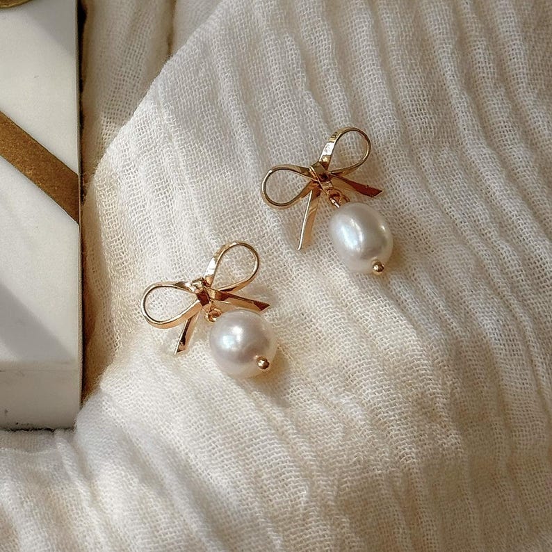 A pair of gold earrings with white pearls and bows, resting on a white fabric surface.