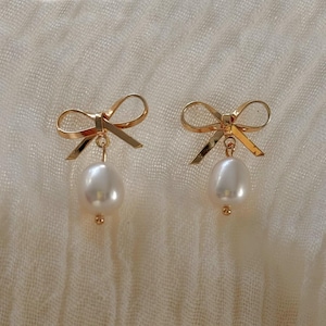 A pair of gold bow earrings with white pearls and bows on a white fabric background.