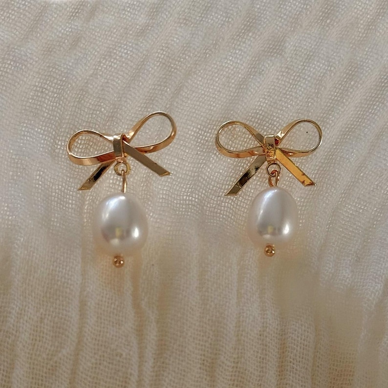 A pair of gold bow earrings with white pearls and bows on a white fabric background.