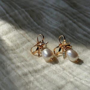 A pair of gold bow earrings with pearl drops laid on a textured light-colored fabric background.