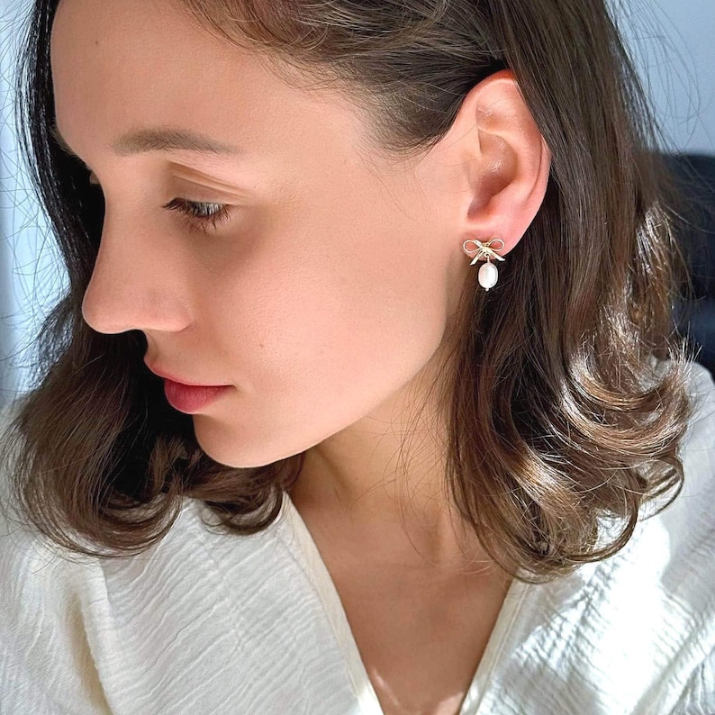 Close-up of a model wearing the earrings, turned in profile. The delicate bow and dangle pearl create an elegant coquette aesthetic for everyday or weddings.
