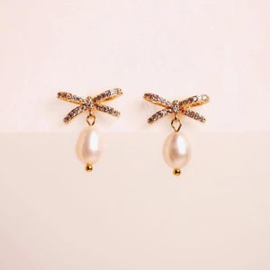 A pair of gold earrings with pearls and rhinestones, displayed against a light pink background.