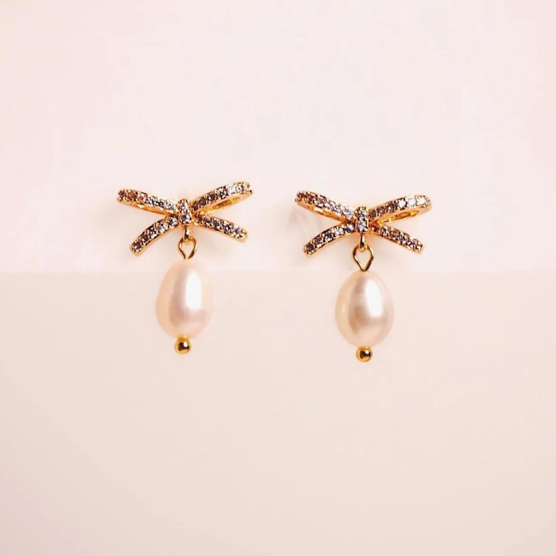 A pair of gold earrings with pearls and rhinestones, displayed against a light pink background.