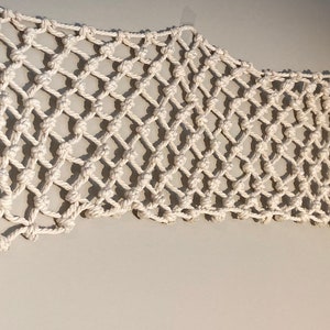 Puede incluir: Un tapiz de macramé blanco con un patrón de diamante. El tapiz está hecho de cuerda gruesa y tiene una franja en la parte inferior.