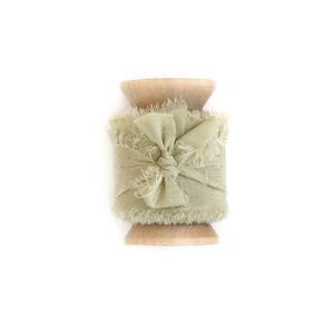 May include: A spool of sage green frayed ribbon with a bow tied in the center. The ribbon is wrapped around a wooden spool.