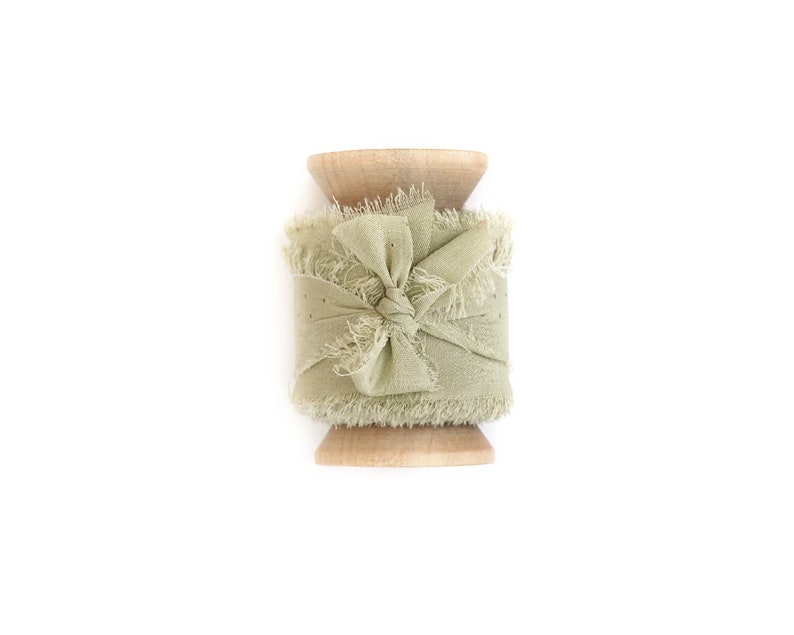 May include: A spool of sage green frayed ribbon with a bow tied in the center. The ribbon is wrapped around a wooden spool.