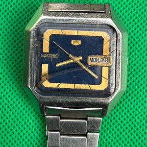Pode incluir: Um relógio de pulso Seiko 5 prateado com caixa quadrada e mostrador azul-escuro com marcadores de hora e ponteiros dourados. O vidro do relógio tem uma rachadura visível, um mostrador de dia e data às três horas e uma pulseira de elos metálicos.