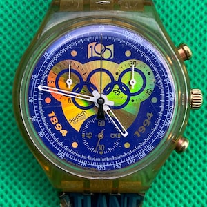 May include: A Swatch chronograph wristwatch with a translucent yellow case and a blue dial featuring the Olympic rings, the years 1894 and 1994, and the text 100. The watch has white hands, three sub-dials, and a blue strap with visible text.