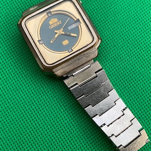 Può includere: Un orologio da polso quadrato color oro con un quadrante circolare verde ottanio scuro che presenta il logo Orient, lancette dorate e una finestrella giorno-data a ore tre. L'orologio include un cinturino a maglie in metallo color argento.