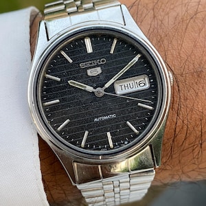 Pode incluir: Um relógio de pulso automático Seiko 5 em tom prateado com um mostrador preto texturizado com linhas horizontais, índices de bastão prateados e uma janela de dia e data na posição das três horas exibindo THU 16. Pulseira de elos metálicos.