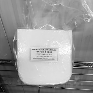 Op de afbeelding: Een blok harde talg, weegt ongeveer 1,13 kg, is verpakt in helder plastic. Het etiket luidt "HARD TALLOW 2.5LBS, BATCH # 1002, PURE: UNBLEACHED, NOT FOR CONSUMPTION, WWW.TENDERLOVINGTALLOW.COM". Het product staat op een metalen plank.
