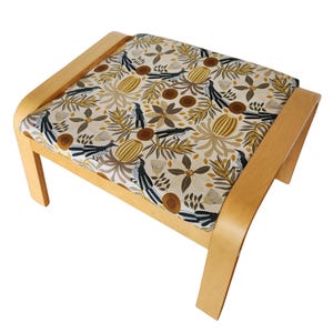 Może przedstawiać: Podnóżek z jasną drewnianą ramą i wzorzystą poduszką. Poduszka ma kremowe tło z powtarzającym się wzorem ptaków, kwiatów i liści w odcieniach brązu, beżu i czerni. Zakrzywione nogi i rama są w kolorze jasnego naturalnego drewna.