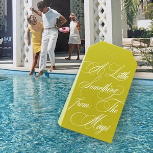 May include: A rectangular chartreuse gift tag with a hole at the top, featuring white cursive text that reads A Little Something From: The Mays, set against a background of a swimming pool with three people standing on the edge of the deck.
