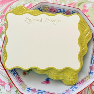 May include: A stack of cream-colored rectangular cards with scalloped edges and a chartreuse border. The text "Kristen & Harrison" is printed in a thin, gold-colored script font at the top center of the top card in the stack.