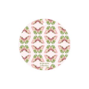 May include: Round ornament with a pink background and a repeating pattern of green and pink butterflies. The text "RAWSON collective" is printed in green at the bottom of the ornament.