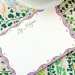 May include: A rectangular stationery card with a white center and a wavy lavender border outlined in gold. The card features small gold and lavender star icons along the border and the name Lila Malone printed in gold cursive script in the upper left.