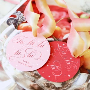 May include: Two circular gift tags with ribbon, one light pink and one red. The pink tag features the text Fa la la la la and LOVE, COCO & DOC in red script. The red tag features the text La la la in white script. Both tags have small star accents.
