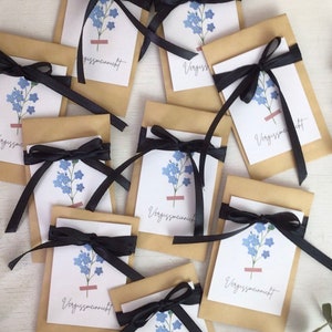 Könnte beinhalten: Eine Sammlung kleiner, brauner Papiertüten, die mit schwarzem Satinband gebunden sind. Jede Tüte hat eine weiße Karte mit einer blauen Blumenillustration und dem Wort "Vergissmeinnicht". Die Tüten sind auf einer weißen Oberfläche angeordnet.