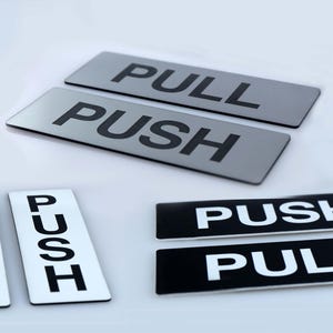 Puede incluir: Un conjunto de letreros rectangulares para puertas con las palabras «TIRAR» y «EMPUJAR» en negrita y mayúsculas. Los letreros están en varios esquemas de color, incluyendo plata con texto negro y blanco con texto negro.