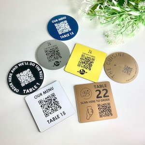 May include: A selection of QR code table signs in different shapes and colours, including blue, yellow, silver, black, and gold. Each sign displays a QR code and text like "Our Menu" and "Table 15".