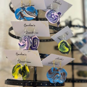 Peut inclure: Assortiment de boucles d'oreilles faites à la main, marbrées et colorées, présentées sur un stand noir. Chaque paire de boucles d'oreilles est sur une carte blanche avec le texte "Cecilia's Sunshine" imprimé dessus.