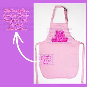 May include: A light pink apron with the text "HOLY SPIRIT ACTIVATED" in a pink and white design. The apron also features a pocket with the quote "Not by might nor by power, but by My Spirit," says The Lord. -Zechariah 4:6."