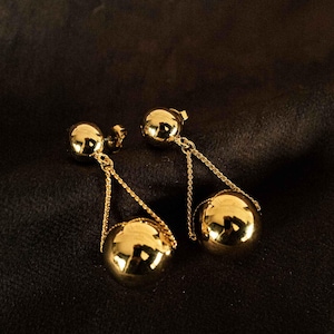 Peut inclure: Une paire de boucles d'oreilles dorées. Chaque boucle d'oreille est composée d'un petit clou et d'une sphère plus grande suspendue, reliée par une fine chaîne. Les sphères reflètent la lumière, créant un aspect brillant. Le fond est une surface sombre et texturée.