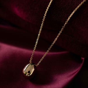 May include: A gold-colored pendant necklace with a teardrop-shaped charm. The charm is smooth and reflective, hanging from a delicate chain. The necklace is displayed against a deep burgundy fabric.