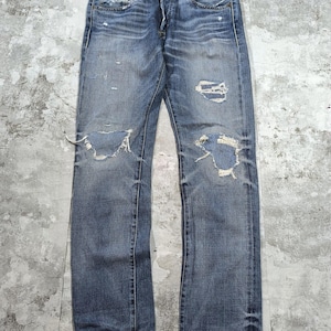 May include: A pair of blue denim jeans featuring a distressed finish with faded whiskering at the hips, a button fly closure, and prominent frayed rips at both knees. The fabric displays a medium wash with lighter highlights and dark seams.