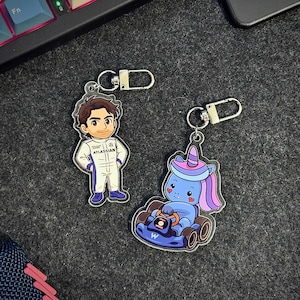 Pode incluir: Dois chaveiros de acrílico. Um apresenta um personagem de desenho animado em um traje de corrida branco com o texto "ATLASSIAN". O outro é um unicórnio azul dirigindo um kart azul. Ambos têm chaveiros prateados.