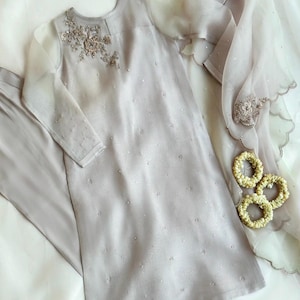 May include: A light gray embroidered kurta with a matching pair of pants and a sheer dupatta. The kurta has a round neckline and long sleeves. The dupatta has a scalloped edge and is decorated with floral embroidery.