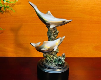 May include: A silver-colored dolphin sculpture featuring two dolphins leaping from a textured, green and brown base. The sculpture sits on a black, round base, and is set against a warm, wooden background.